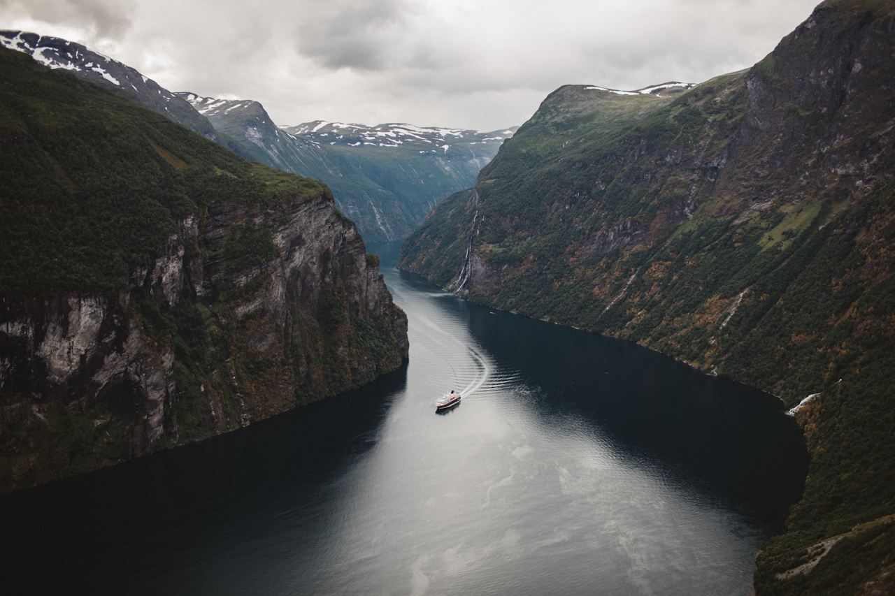 Aerial editorial photograph of a single ship navigating a Nordic fjord between towering cliffs.